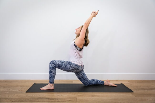 woman doing a low lunge yoga pose with arms raised on a grey mat